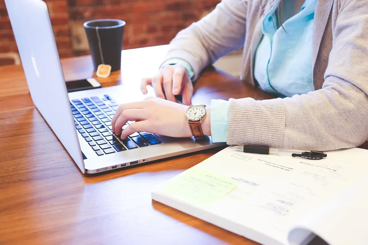 Une personne tape sur le clavier d'un ordinateur portable posé sur un bureau en bois, à côté d'un cahier ouvert et d'un gobelet de café.