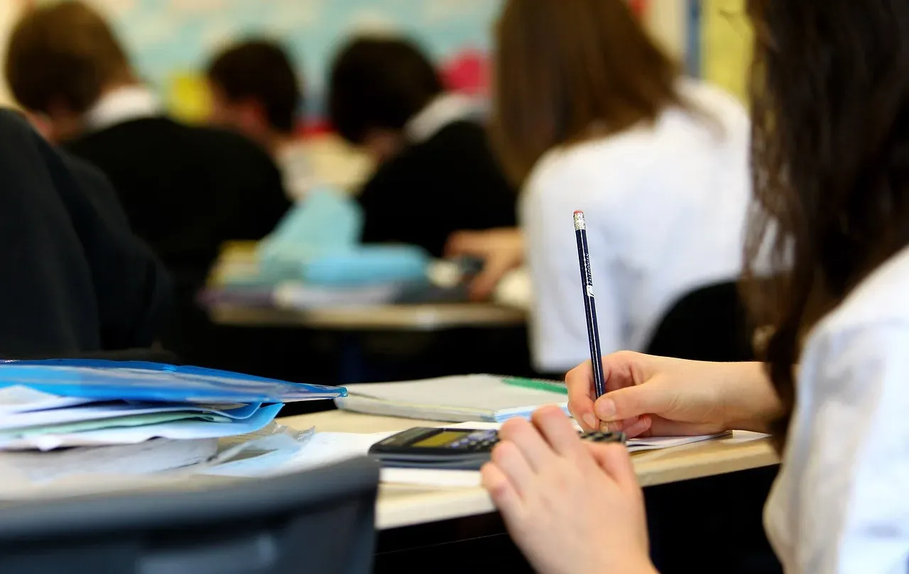 Des élèves assis à leurs tables dans une salle de classe, cahiers ouverts, l'un d'eux en train d'écrire au crayon.