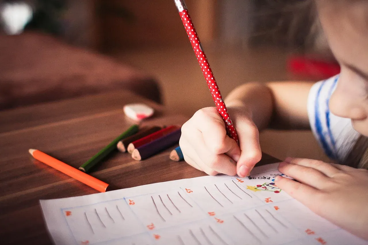 Gros plan sur la main d'un enfant qui écrit au crayon rouge sur une feuille d'exercices, des crayons de couleur éparpillés sur la table en bois.