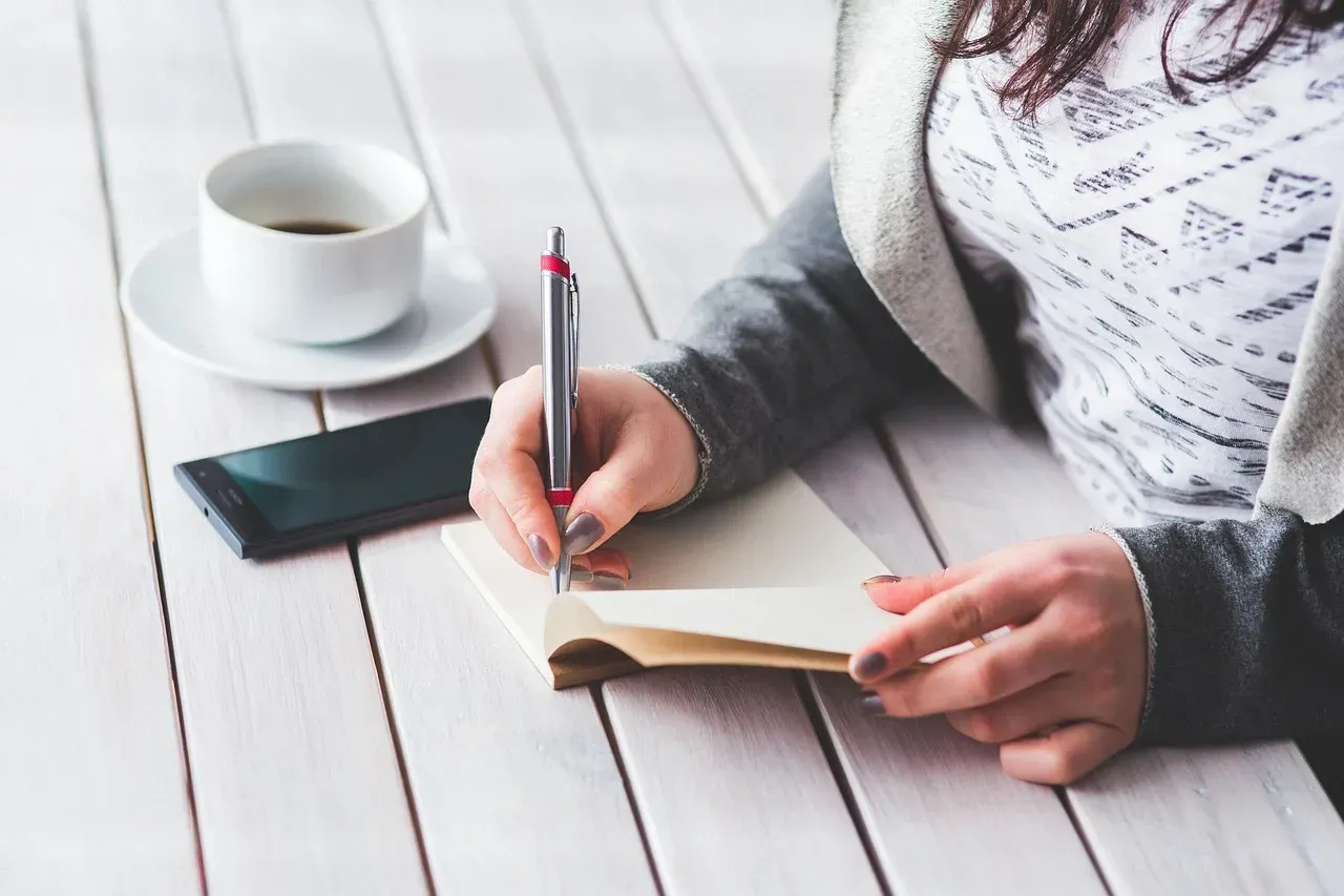 Une jeune femme écrit dans un carnet posé sur une table en bois, à côté d'une tasse de café et d'un téléphone.