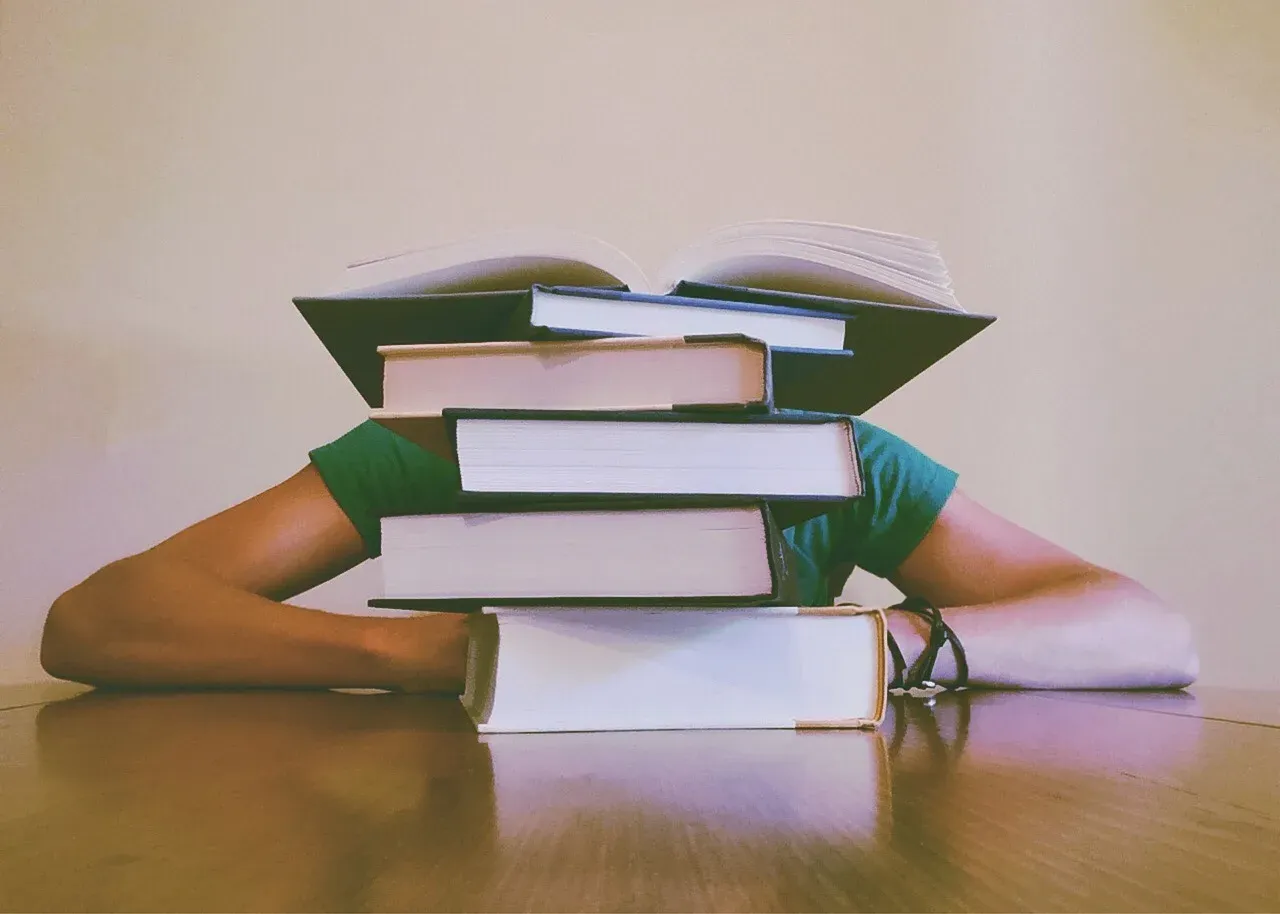 Une personne assise à une table, le visage entièrement caché derrière une pile de livres empilés devant elle.