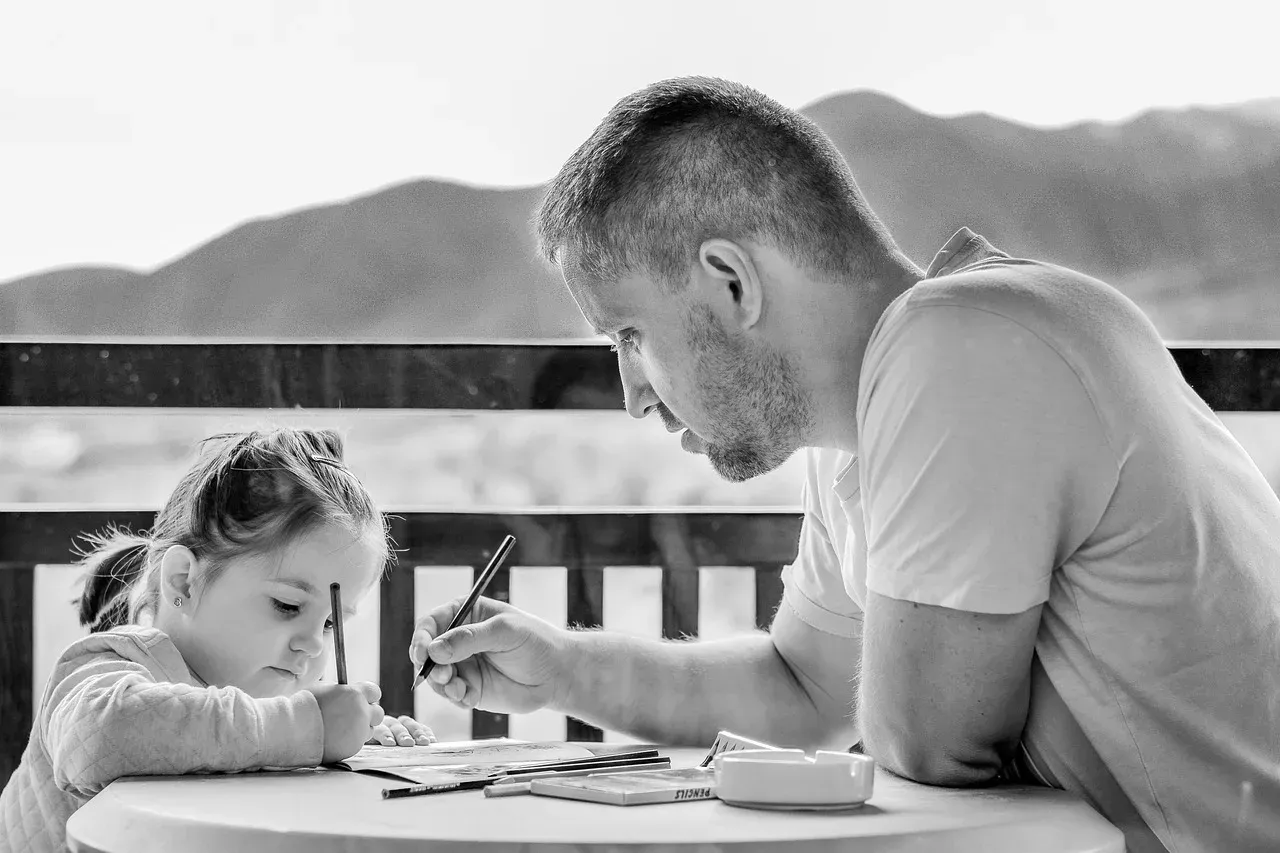 Photographie en noir et blanc d'un adulte et d'un enfant assis à la même table, penchés chacun sur une feuille, en train d'écrire.