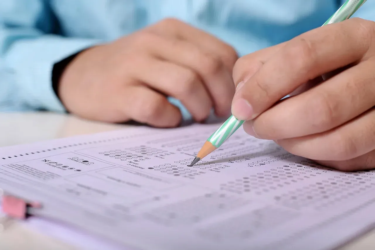 Gros plan sur une main qui remplit au crayon à papier une grille de réponses à cases, sur une table d'examen.