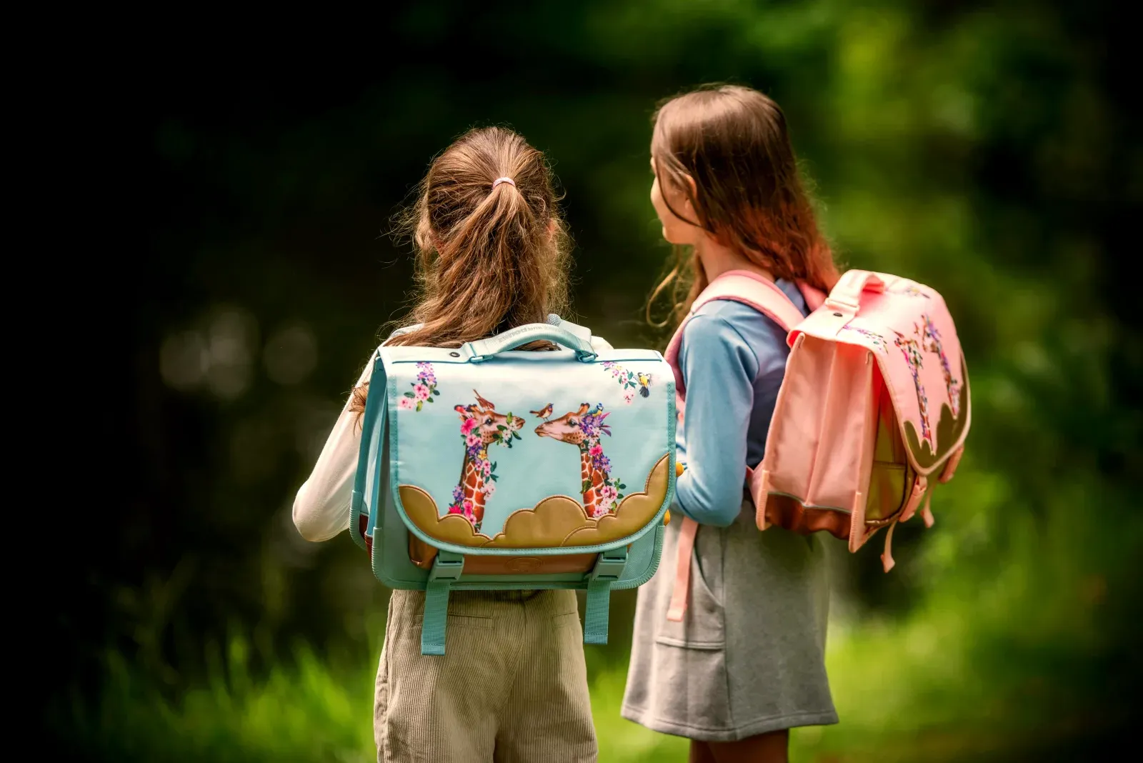 Deux jeunes filles de dos, portant des cartables colorés illustrés de girafes, prêtes pour l'école dans un décor verdoyant.
