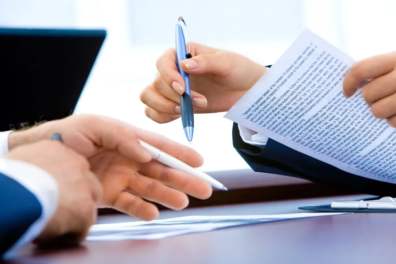 Gros plan sur des mains en costume : l'une tend un stylo au-dessus d'un document imprimé, un ordinateur portable ouvert derrière.