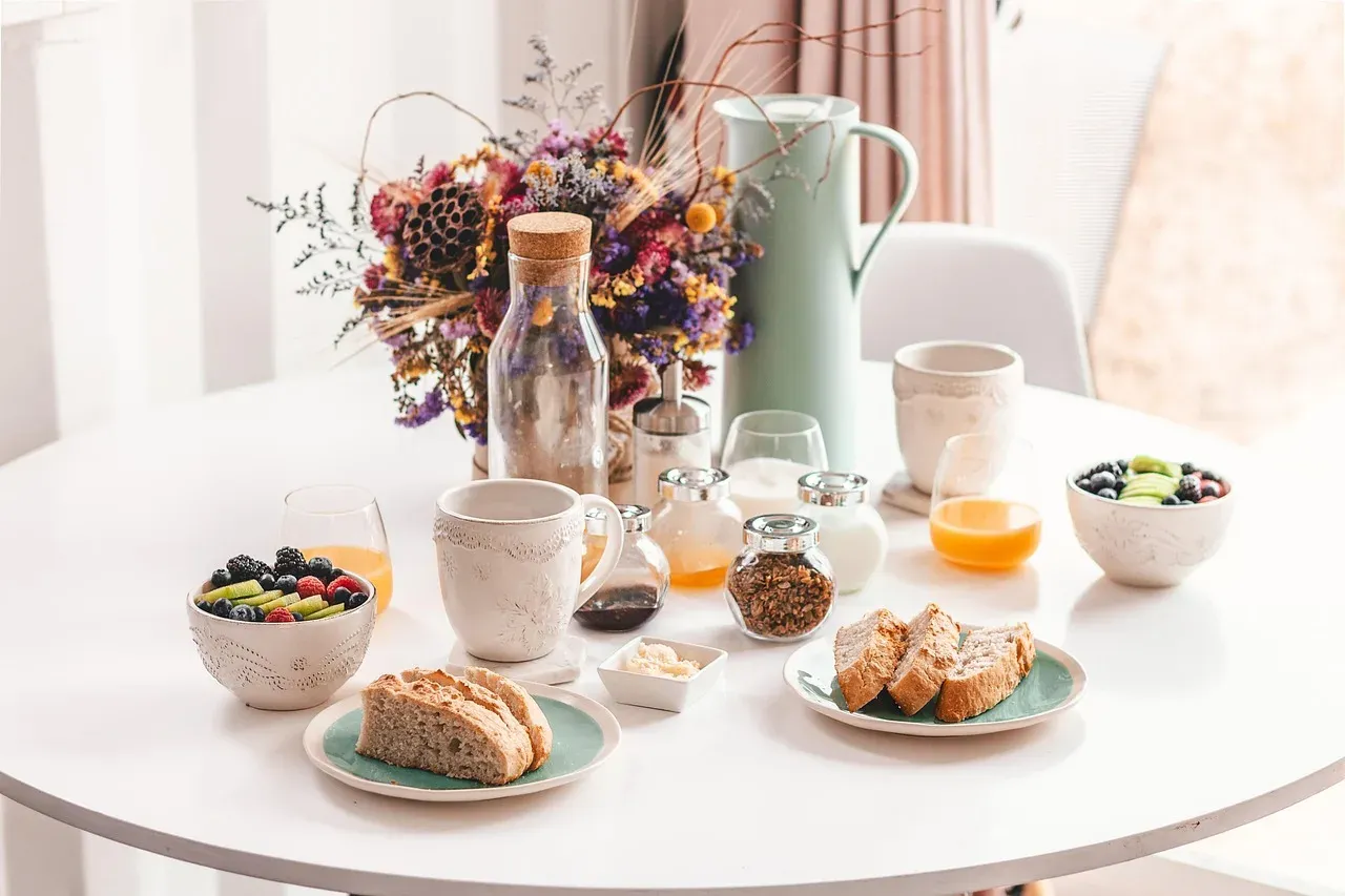 Une table ronde dressée pour le petit-déjeuner : pain, fruits rouges, jus d'orange, bols et pichet, avec un bouquet de fleurs.