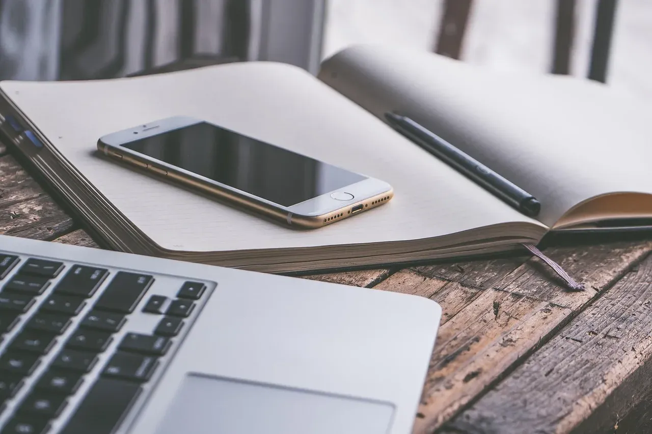 Un ordinateur portable, un smartphone et un carnet ouvert avec un stylo, posés côte à côte sur un bureau en bois.