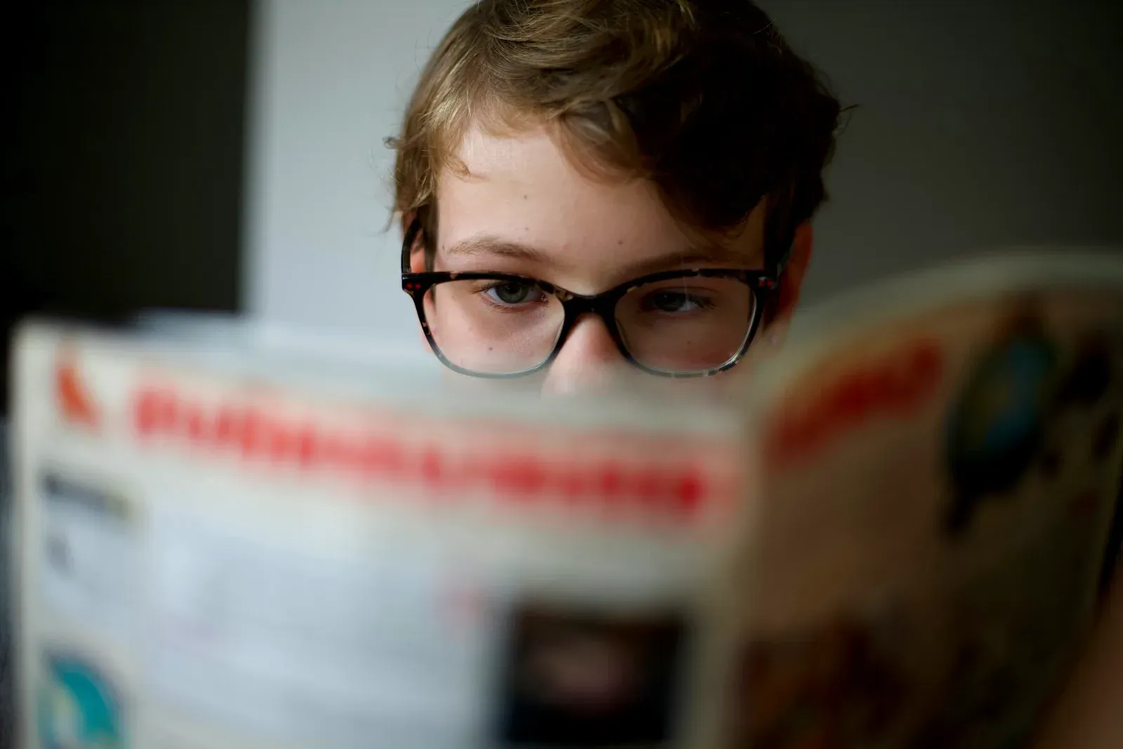 Jeune garçon portant des lunettes à monture écaille, concentré sur la lecture d'un livre ouvert tenu près de lui, le premier plan étant flou.