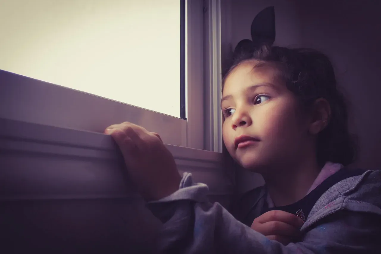 Une fillette en sweat à capuche, accoudée au rebord d'une fenêtre, le regard tourné vers l'extérieur, l'air songeur.